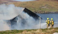 Two firefighters in gear spray water on a smoking building near a body of water in a grassy, hilly area.