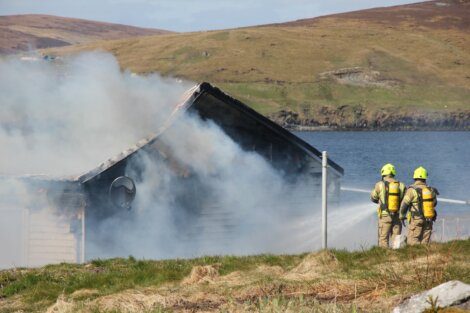 Two firefighters in gear spray water on a smoking building near a body of water in a grassy, hilly area.