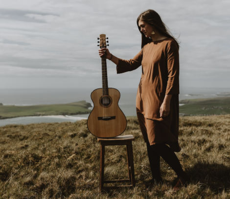 A woman in a brown dress stands on a grassy hill, holding an acoustic guitar by its neck, with a wooden stool next to her. A coastal landscape with cloudy skies is visible in the background.