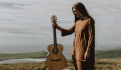 A person in a brown dress stands outdoors on a hill, holding a guitar upright. The background shows a coastline with fields and a cloudy sky.