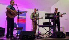 Three men play guitars and a banjo on stage in front of microphones and speakers, with a purple backdrop and a banner listing insurance and financial services.