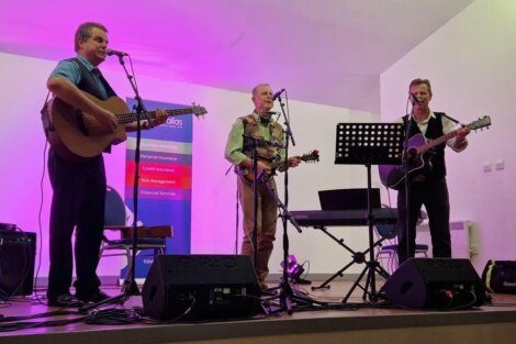 Three men play guitars and a banjo on stage in front of microphones and speakers, with a purple backdrop and a banner listing insurance and financial services.