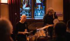 Two musicians perform on stage, one playing a drum set and the other playing a handheld drum, in front of a stained glass window.