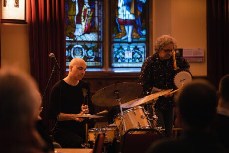 Two musicians perform on stage, one playing a drum set and the other playing a handheld drum, in front of a stained glass window.