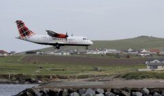 A Loganair aircraft is seen landing on a runway near the coast with houses and grassy hills in the background.