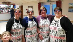 Four people wearing aprons with various texts and novelty glasses stand together in a room. A child is smiling in the foreground. A bar and another person are visible in the background.