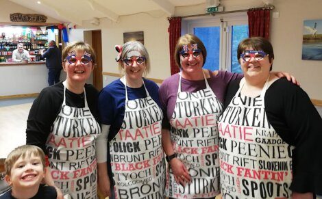 Four people wearing aprons with various texts and novelty glasses stand together in a room. A child is smiling in the foreground. A bar and another person are visible in the background.