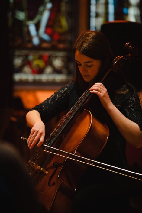 A woman with long brown hair plays a cello in a dimly lit room with stained glass windows in the background.