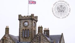 A stone building with a clock tower and a British flag on top. An emblem in the corner reads "King Charles III Coronation 6th May 2023".