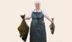 A person wearing a striped apron and a hairnet holds a large fish in each hand, standing against a plain background.