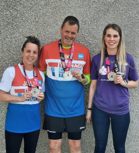 Three people standing in front of a gray wall, each holding a medal around their necks. They are dressed in athletic wear and are smiling at the camera.