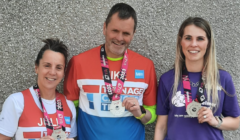 Three people wearing race medals and branded shirts stand together and smile at a wall.