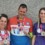 Three people wearing race medals and branded shirts stand together and smile at a wall.