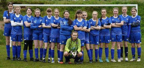 A girls' soccer team wearing blue uniforms poses on a field in front of a goal. One player wearing a green goalkeeper jersey kneels in front, while the others stand with arms crossed.