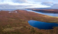 Aerial view of a vast, brown-colored landscape with a large blue lake and a smaller waterbody, bordered by hills and a distant coastline under a partly cloudy sky.