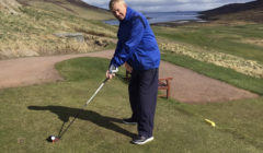 A person in a blue jacket and dark pants is preparing to swing a golf club on a golf course with rolling hills and a body of water in the background.