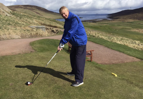 A person in a blue jacket and dark pants is preparing to swing a golf club on a golf course with rolling hills and a body of water in the background.