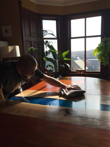 A person with a bald head, beard, and tattoos polishes a wooden table in a room with large windows and potted plants. Sunlight streams through the windows, illuminating the area.