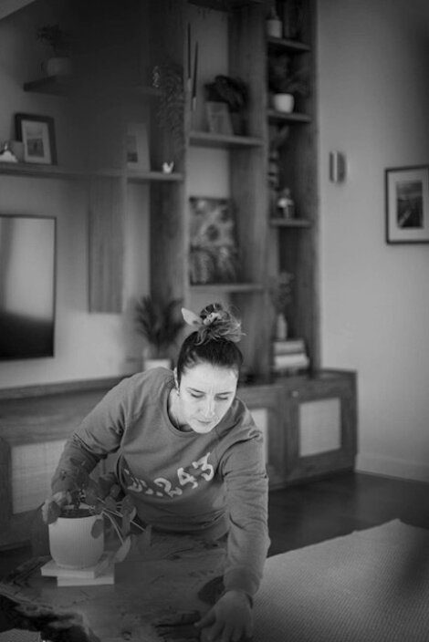 A woman in a casual outfit arranges items on a tabletop in a living room with a shelving unit in the background. The image is in black and white.