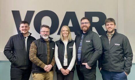 Five smiling individuals pose in front of a wall with the letters "VOAR." They are all wearing black jackets with the same letters printed on them.