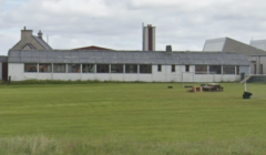 A long, single-story building with a corrugated metal roof, surrounded by a grassy field. Additional buildings with chimneys are visible in the background.