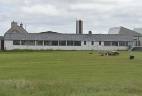 A long, single-story building with a corrugated metal roof, surrounded by a grassy field. Additional buildings with chimneys are visible in the background.