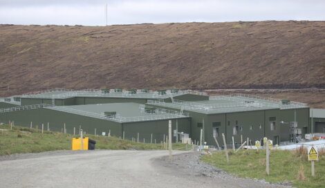 A large, green industrial building with a flat roof in a rural area surrounded by a gravel road and grassy field.