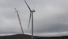 A large crane works beside a wind turbine under a cloudy sky on a barren landscape.