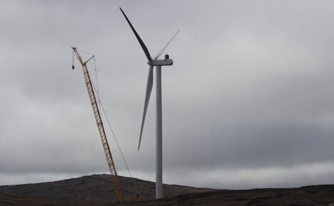 A large crane works beside a wind turbine under a cloudy sky on a barren landscape.