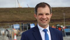 A man in a blue suit and tie smiles at the camera, standing outdoors with construction equipment and a hilly landscape in the background.