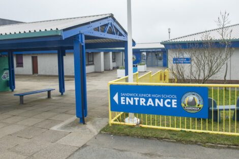 Entrance sign for Sandwick Junior High School, featuring a blue and yellow color scheme. The school building is visible in the background.