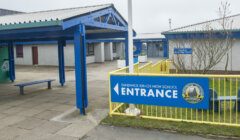 Entrance sign for Sandwick Junior High School, featuring a blue and yellow color scheme. The school building is visible in the background.