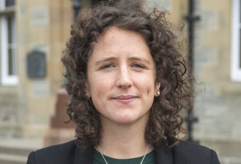 A person with curly hair and a slight smile stands in front of a building wearing a dark blazer and green shirt.