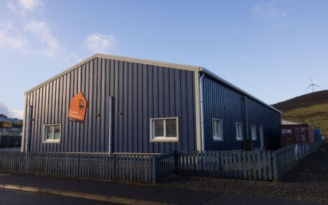 A blue metal building with a sign for a local food bank stands on a gray pavement, surrounded by a wooden fence. A wind turbine is visible on a hill in the background.