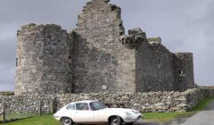 A vintage white car is parked on a road in front of an old stone castle with two round towers and a partially cloudy sky in the background.