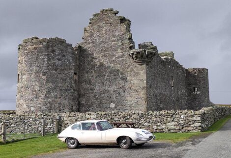 A vintage white car is parked on a road in front of an old stone castle with two round towers and a partially cloudy sky in the background.