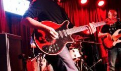 A guitarist performs on stage with a red electric guitar, accompanied by a bassist and drummer, under red stage lighting.