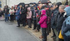 A group of people, some holding umbrellas and wearing rain gear, stand on a sidewalk in wet weather, observing an outdoor event. Some hold small flags and flowers.