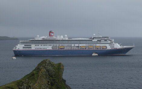 A large blue and white cruise ship with "Fred. Olsen Cruise Lines" on the side sails in the ocean, with a rocky outcrop in the foreground and overcast skies in the background.