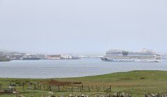 Several cruise ships are docked near a port on a foggy day. A grassy field and stone fence are in the foreground.