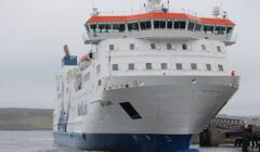 A large white and blue ferry is docked at a pier on a cloudy day, with hills visible in the background.