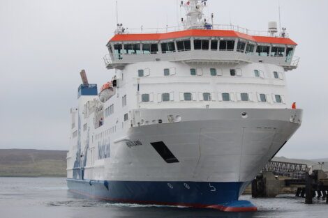 A large white and blue ferry is docked at a pier on a cloudy day, with hills visible in the background.