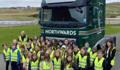 A large group of children and two adults, all wearing high-visibility vests, stand in front of a green truck labeled "NORTHWARDS" and "SHETLAND" in an outdoor area.