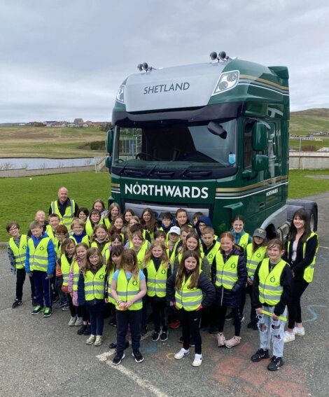 A large group of children and two adults, all wearing high-visibility vests, stand in front of a green truck labeled "NORTHWARDS" and "SHETLAND" in an outdoor area.