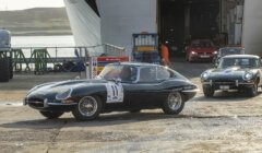 Two vintage sports cars drive off a ferry. The lead car has a racing number on the door, with another similar car following behind. The background shows the open ferry ramp and industrial surroundings.