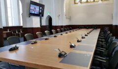 A conference room with a long wooden table and multiple chairs. Microphones are set up at each seat. A video conference is displayed on a wall-mounted screen.