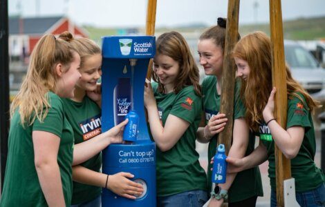 Five young women in green shirts fill blue water bottles at a Scottish Water refill station outdoors.