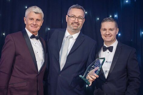 Three men in formal attire stand together. The man in the middle holds a glass triangular award.