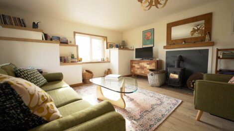 A cozy living room with a green sofa, glass coffee table, wood burning stove, and books on built-in shelves. A wall mirror and television are also visible. Natural light enters through the window.