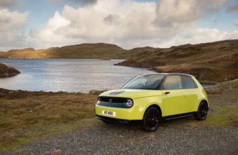 A yellow car is parked on a gravel path beside a large body of water with rolling hills in the background under a cloudy sky.